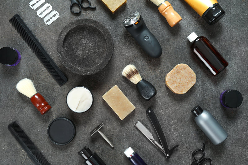 Laid out on a table are shaving items such as razors, shaving cream, and shaving cream brushes for toiletry bags