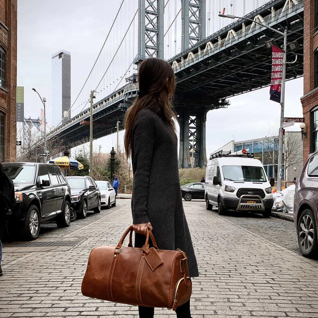 Elegant lady in dark long coat on brick road holding a Rushmore Weekender Ov ernight Duffle Bag with bridge in the background