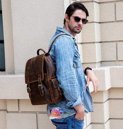 Man in Blue Jean denim jacket leaning against a white granite wall is carrying a Stonewall Leather Backpack over his shoulder